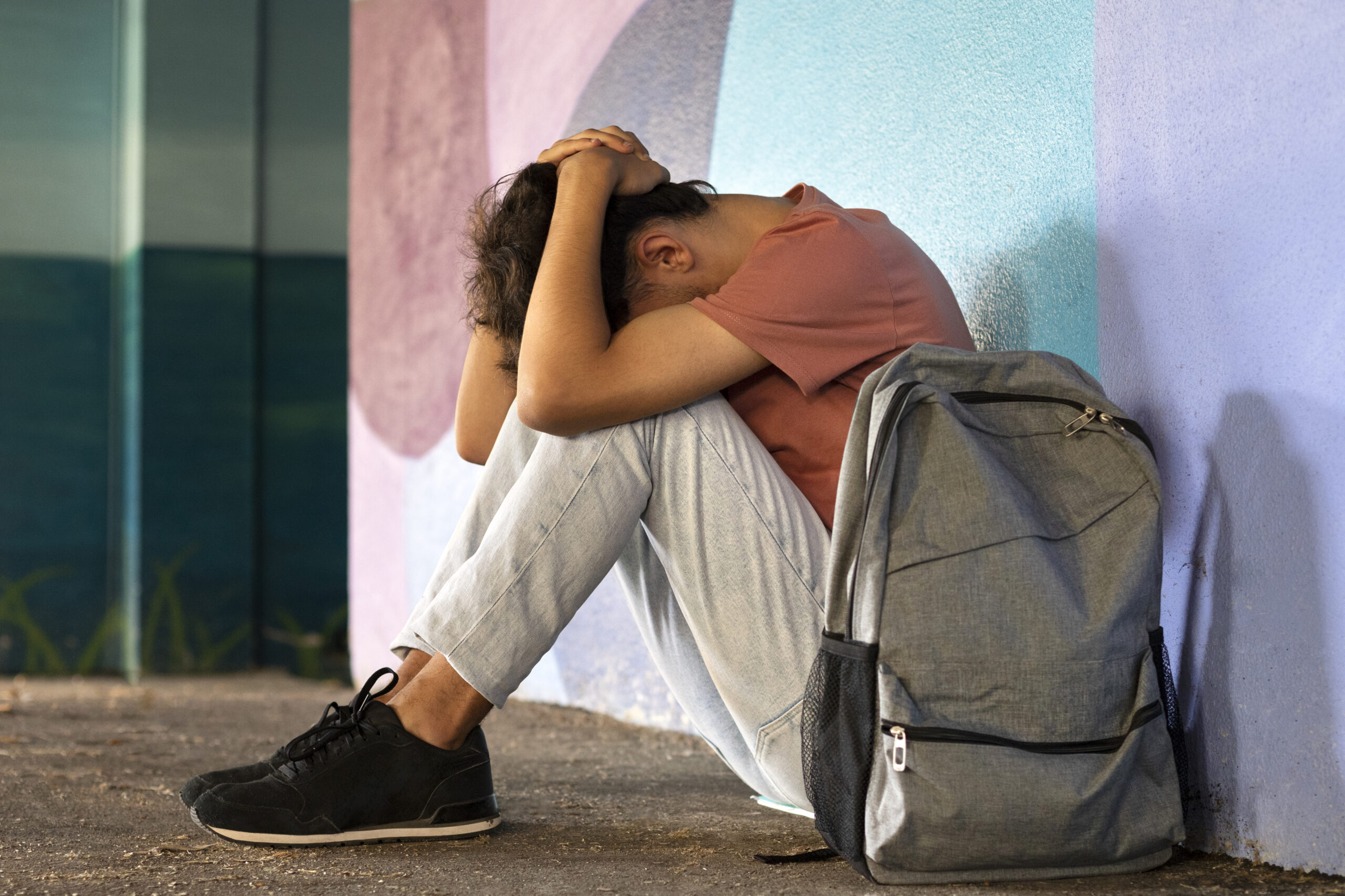 Um estudante sentada no chão e encostada na parede com as mãos sobre a cabeça baixa com uma mochila ao seu lado aparentando estar sofrendo bullying nas escolas.